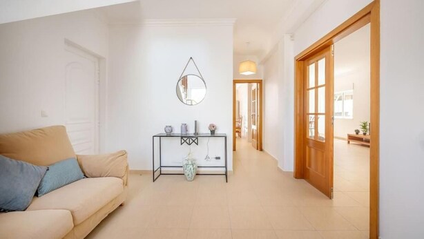 En moderne stue med en beige sofa, et konsolbord med dekorative genstande, et spejl og en trædør, der fører til et andet rum.