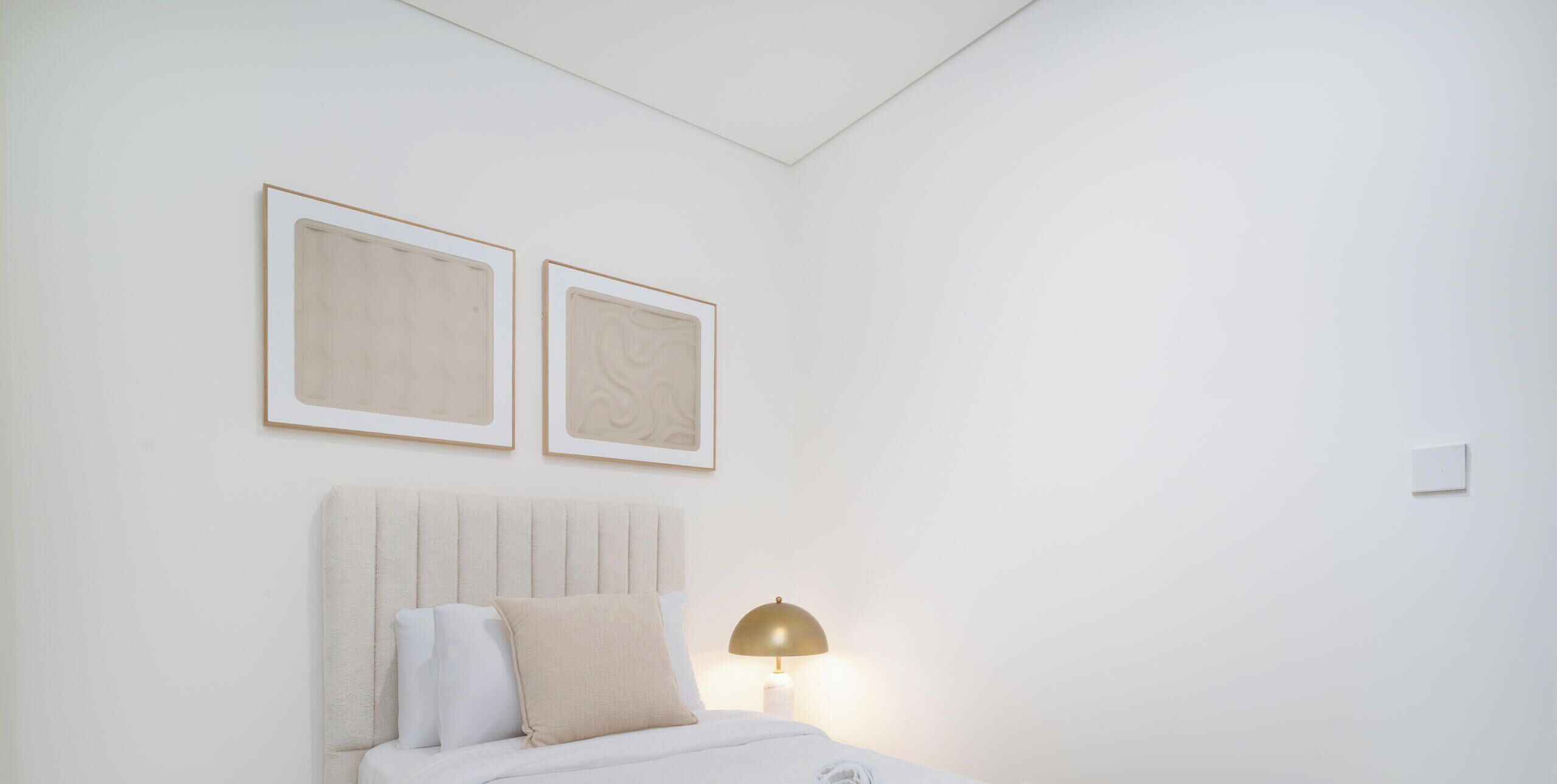 Un dormitorio ordenado con una cama, almohadas, una lámpara y dos cuadros en la pared.
