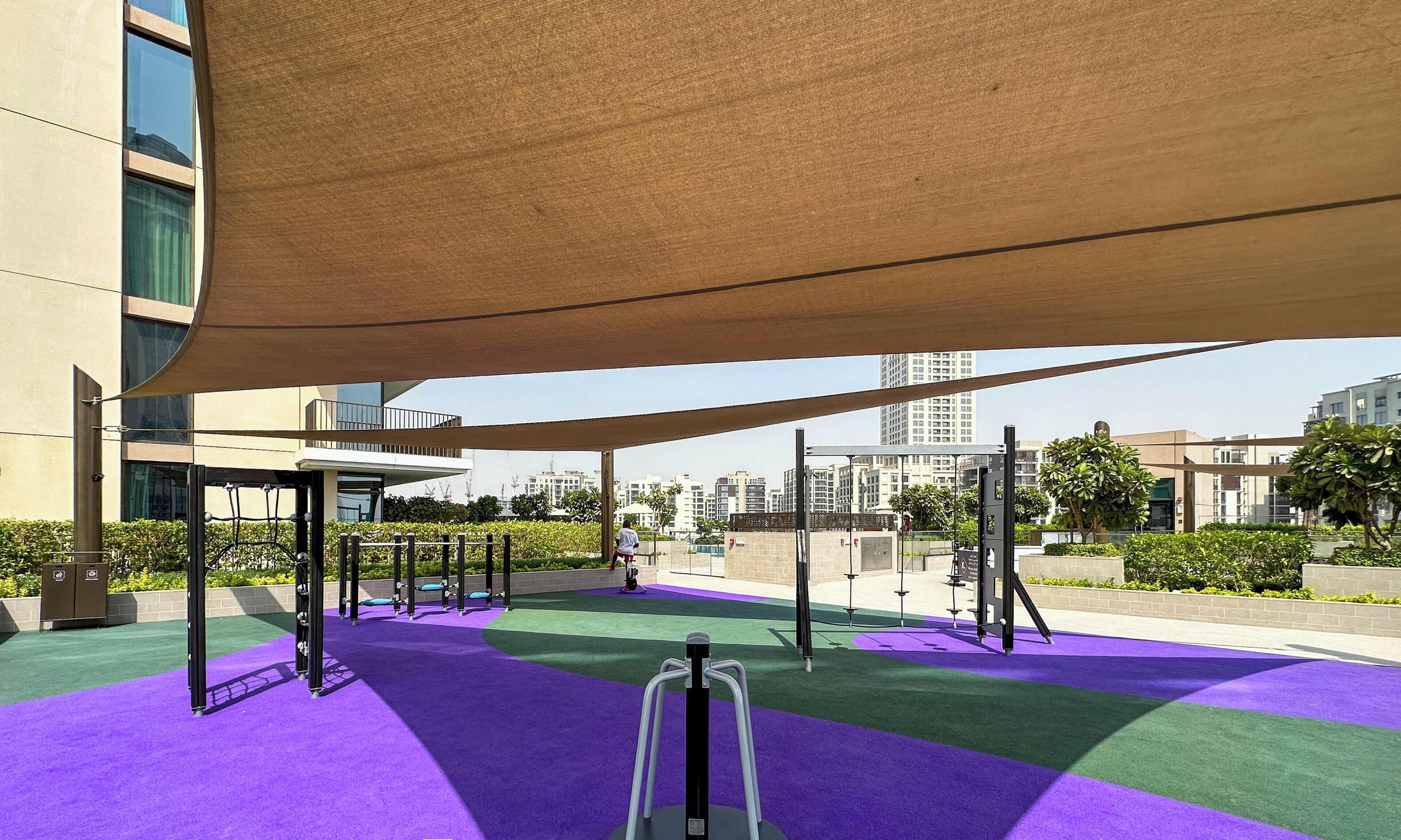 Un patio de juegos en la azotea con equipo de ejercicio y una zona techada, rodeado de edificios y vegetación.