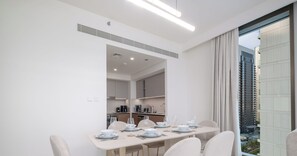 Een moderne eethoek met een lange tafel voor zes personen, witte stoelen en een uitzicht op een stadslandschap door grote ramen.
