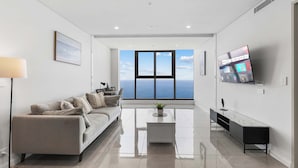 A modern living room with a sofa, a coffee table, a TV, and a large window offering a view of the sky and sea. - 『CASSA OCEAN』Skyline Oceanfront 2BR in Surfers "M" (Surfers Paradise)
