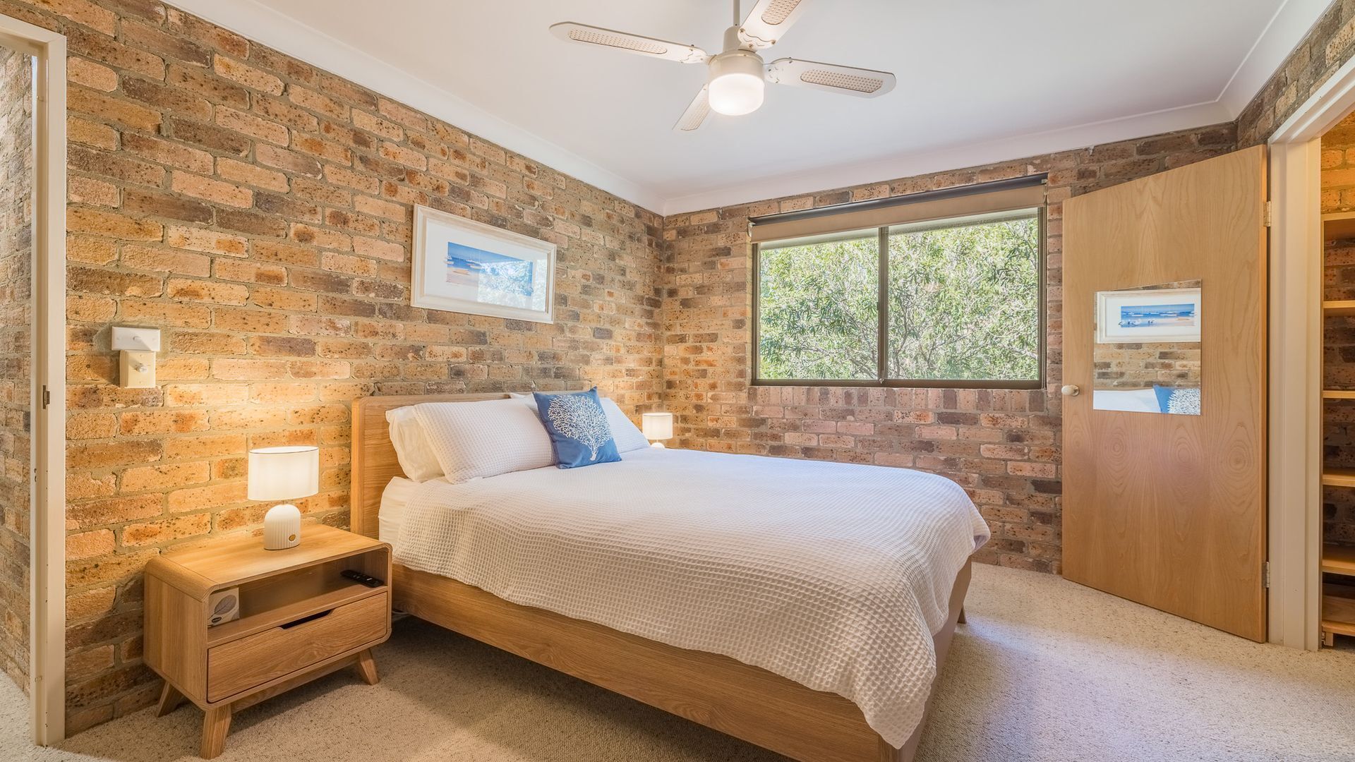 Ein Schlafzimmer mit Backsteinwand, einem Bett mit Holzbettgestell, einem Nachttisch, einer Nachttruhe, einem Deckenventilator und einem Fenster mit Blick auf Grünflächen.