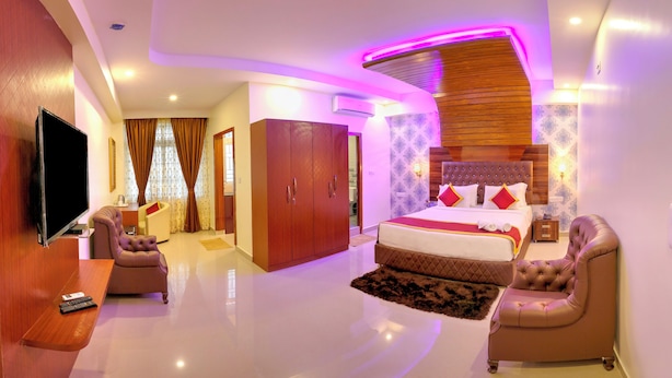 A modern hotel room with a large bed, a sitting area with a sofa and armchair, a flat-screen TV, and a wooden headboard. - Hotel The Prince Royal  (Bengaluru)