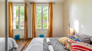 A bedroom with two beds, a window with curtains, a small blue stool, and a bedside table with a lamp.