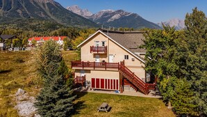 A two-story house with a red balcony and wooden staircase, surrounded by trees and mountains in the background. - 5 Mins from the Resort | Sleeps 3 | Dog Friendly (Fernie)