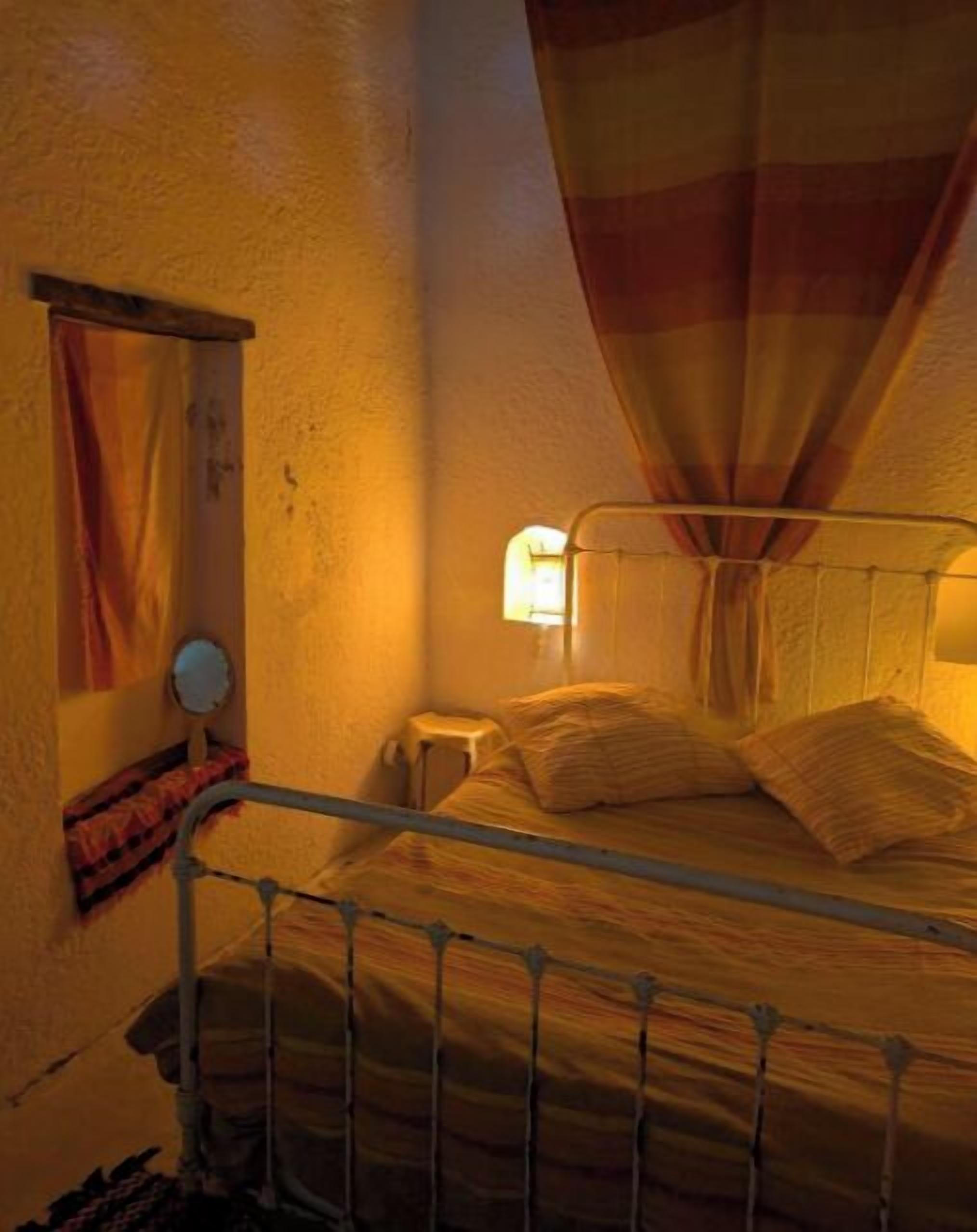 Un lit simple avec un cadre en métal, une table de chevet, une lampe et un rideau.