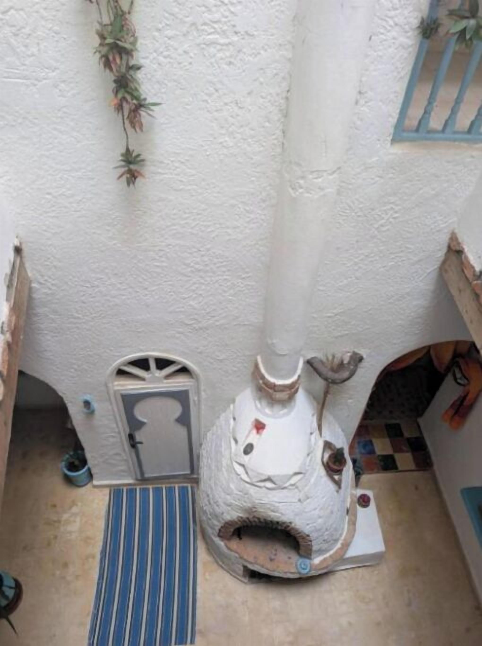 Une pièce de taille modeste avec un mur blanc à texture, une petite porte, un tapis à rayures bleues et un poêle blanc doté d’une cheminée.