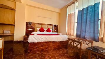Habitación de hotel con piso de parquet de madera, cama con almohadas rojas, cabecera de madera, una mesita de madera y una ventana con cortinas.
