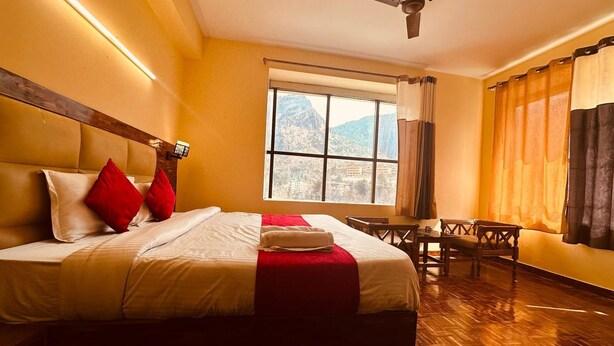 A hotel room with a large bed, a ceiling fan, a window with a mountain view, and a wooden floor. - The Kanchanikoot (Manali)