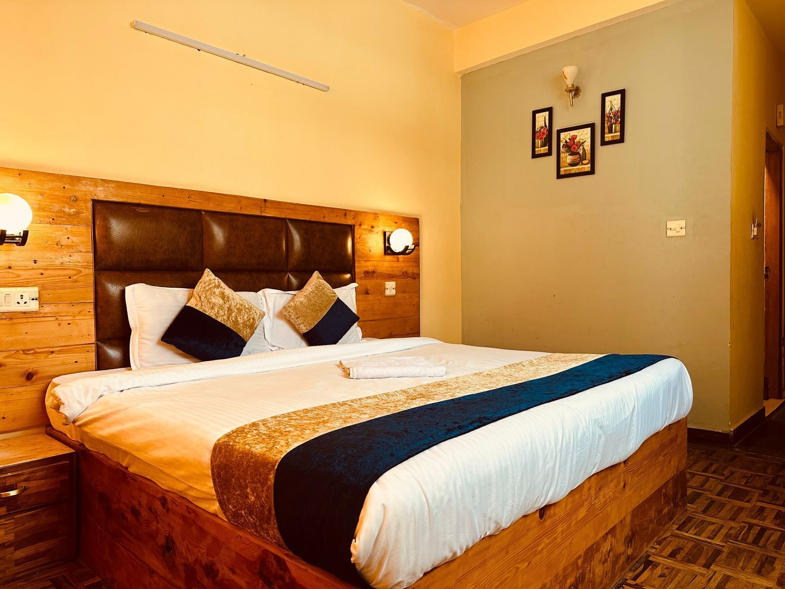 A hotel room with a wooden headboard, a large bed with white and dark pillows, a blue and white throw blanket, and a wall-mounted light fixture.