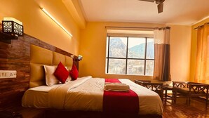 A hotel room with a large bed, wooden headboard, a view of mountains through the window, a ceiling fan, and a table with chairs. - The Kanchanikoot (Manali)