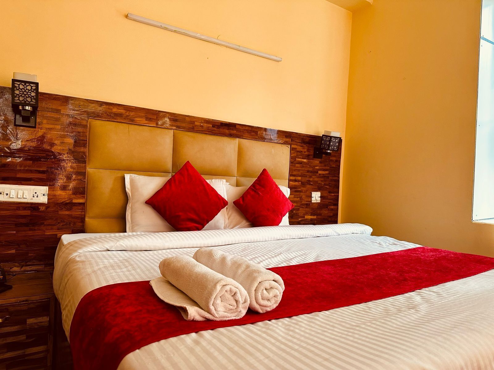 Una habitación de hotel con cabecera de madera, una colcha blanca y roja, y dos toallas enrolladas sobre la cama.