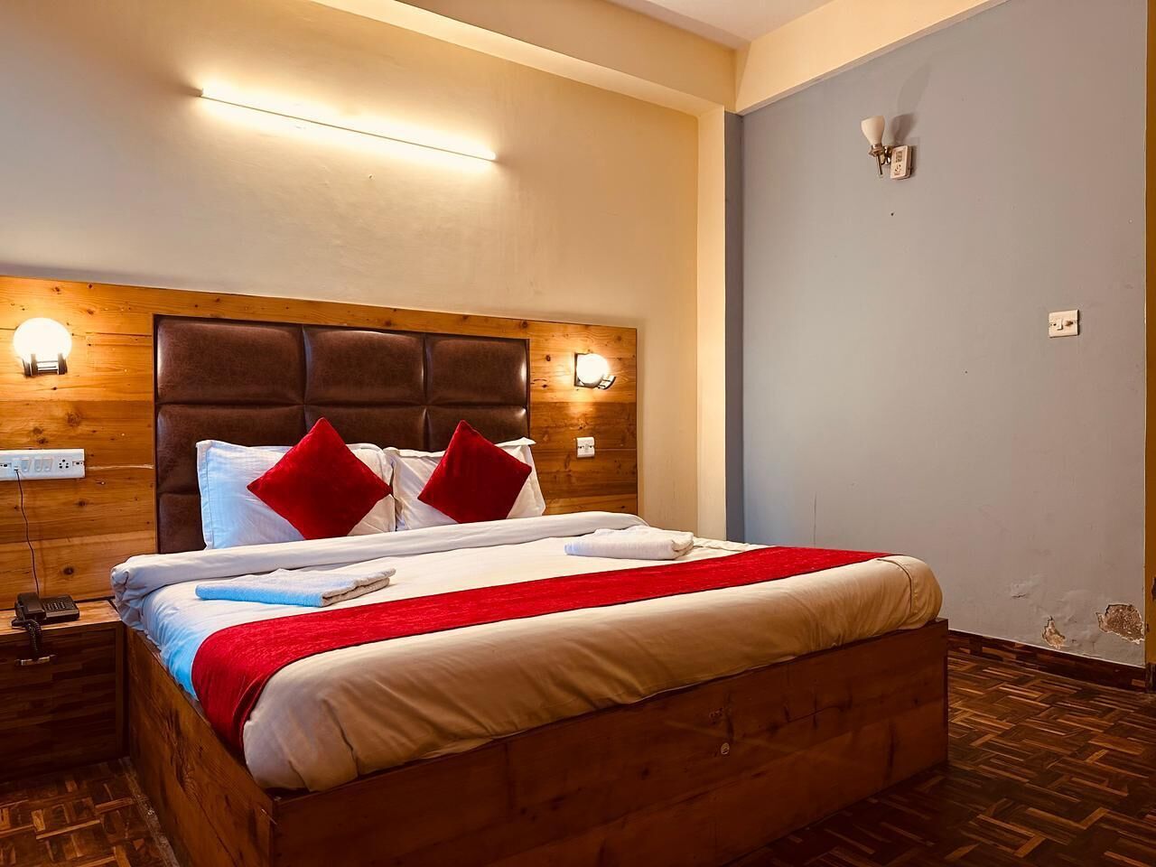 Un lit bien fait, avec une tête de lit en bois, une literie blanche et des oreillers rouges. Deux appliques murales sont fixées de part et d’autre du lit.