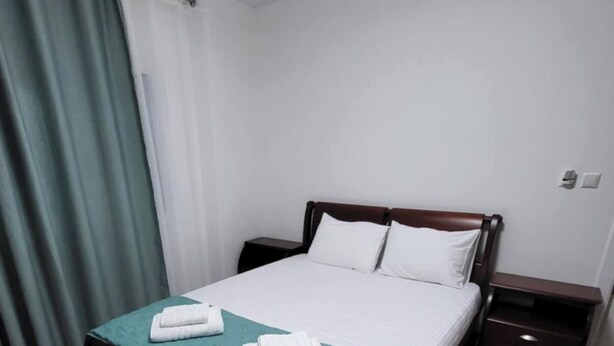 A neatly made bed with a teal bedspread and two white towels, a wooden headboard, a nightstand with a drawer, and a curtain on the left. - Lakeside View Apartment (Udënisht)