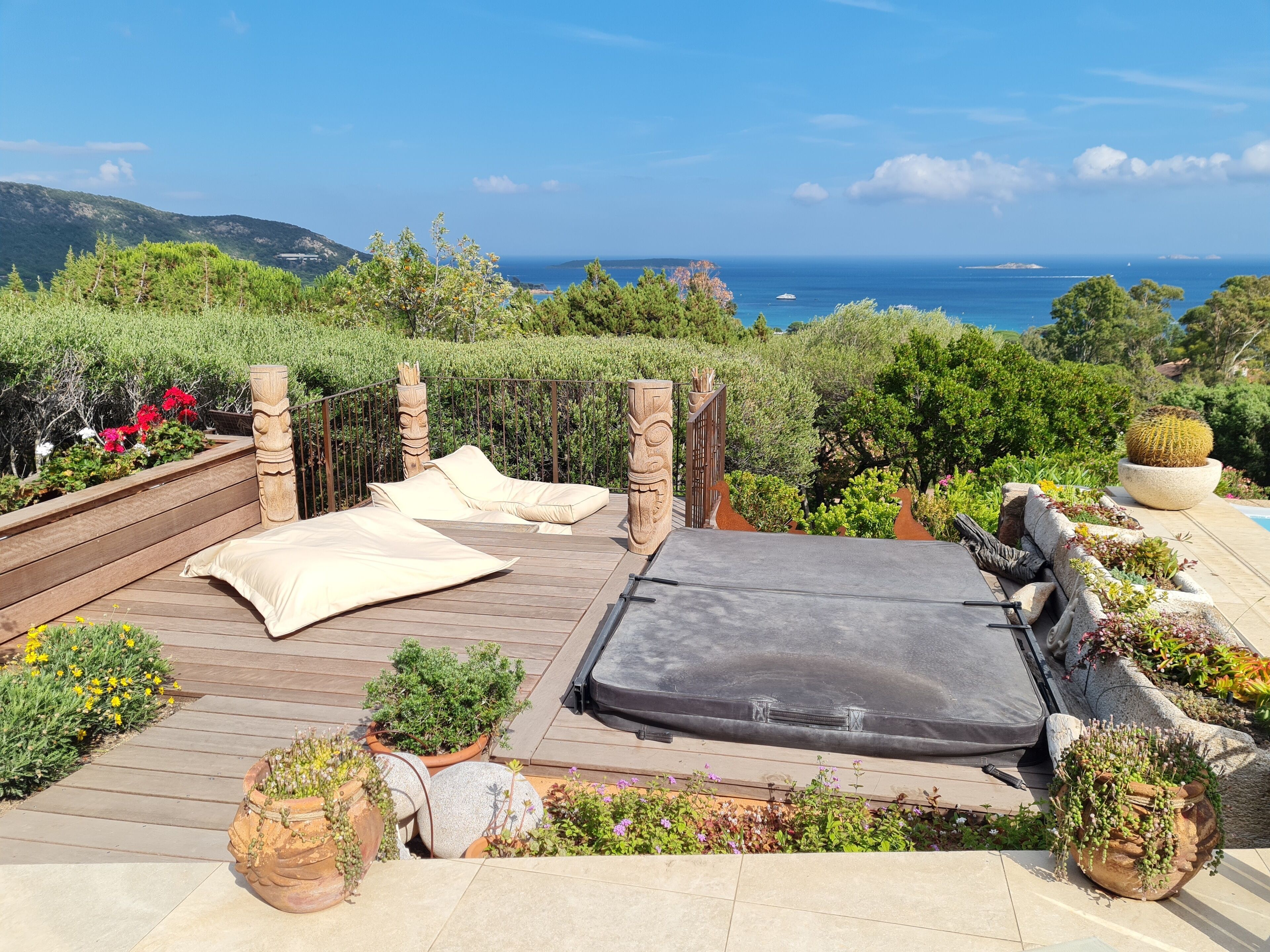 Eine sonnendurchflutete Terrass mit einer Couch und einer Matratze, umgeben von Topfpflanzen und Blick auf das Meer.