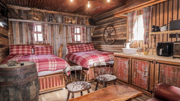 A rustic cabin interior with two beds, a wooden table, leather chairs, a bar area, and a kitchen with a sink and appliances. - Rusty Barn (Limavady)