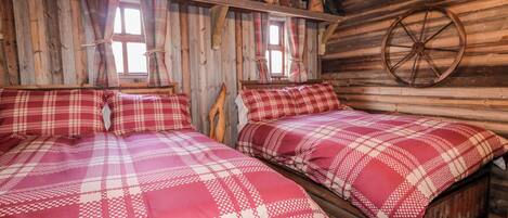 Ein rustikales Holzhütten-Zimmer mit zwei Betten, Karo-Bettwäsche, einem Holzwagenrad und einem Fenster mit Vorhängen.
