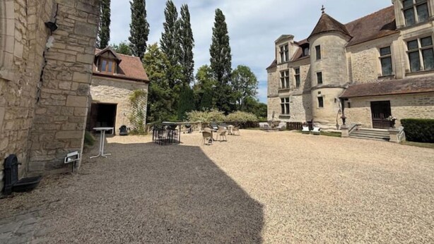 Une cour bordée de bâtiments en pierre, un chemin de gravier et des espaces de repos aménagés à l’extérieur.