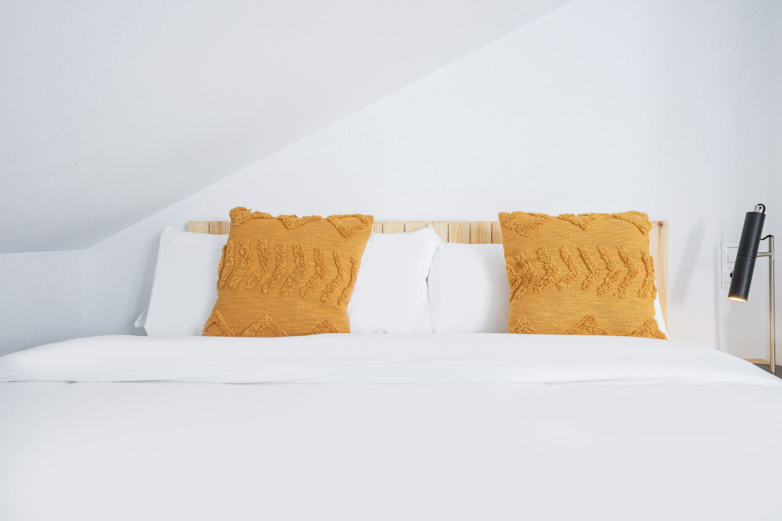 A neatly made bed with white and mustard-colored pillows, a wooden headboard, and a black floor lamp on a white wall.