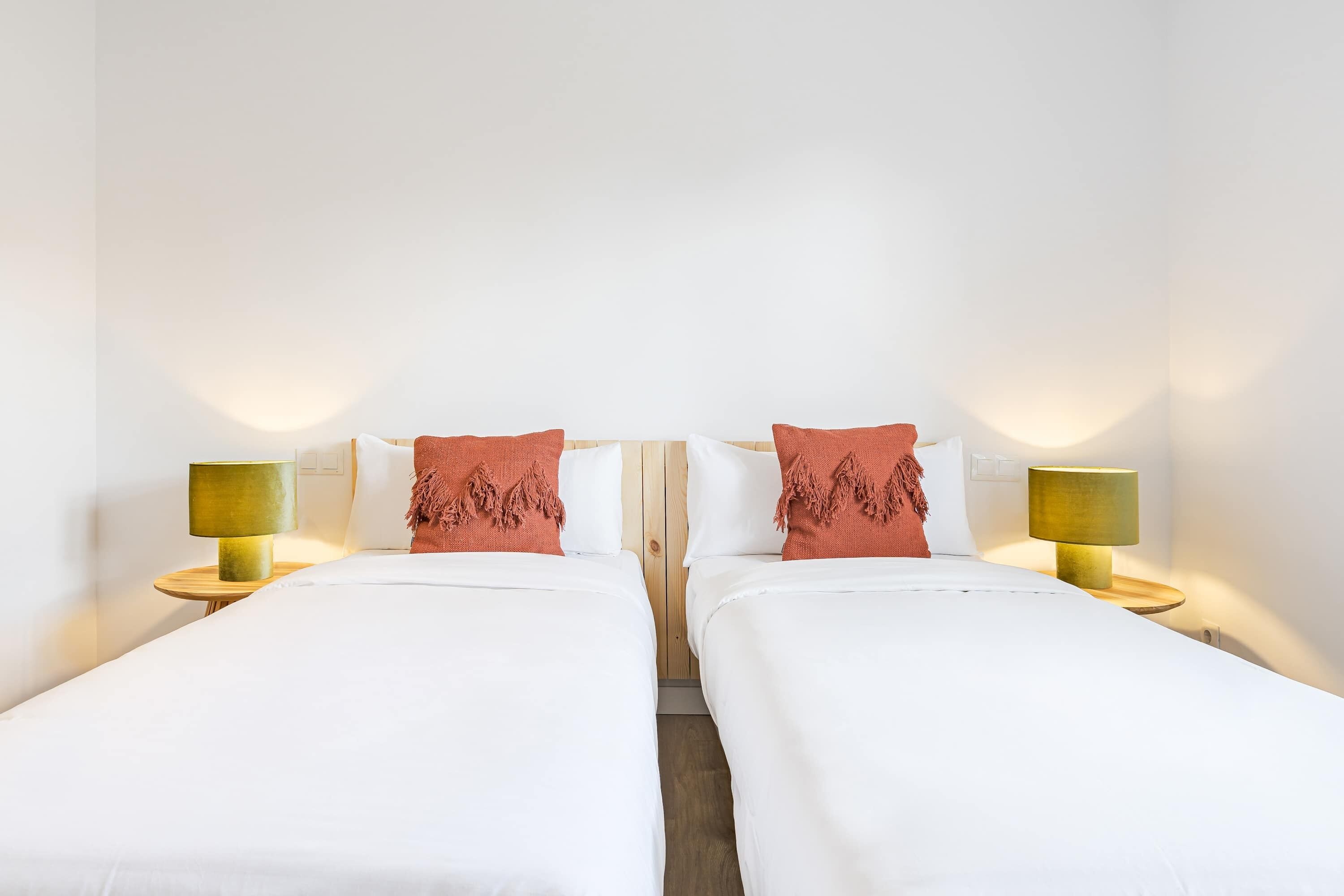 A hotel room with two single beds, each with a wooden headboard, white bedding, and a red throw pillow. There are two bedside tables with lamps, and the walls are white.
