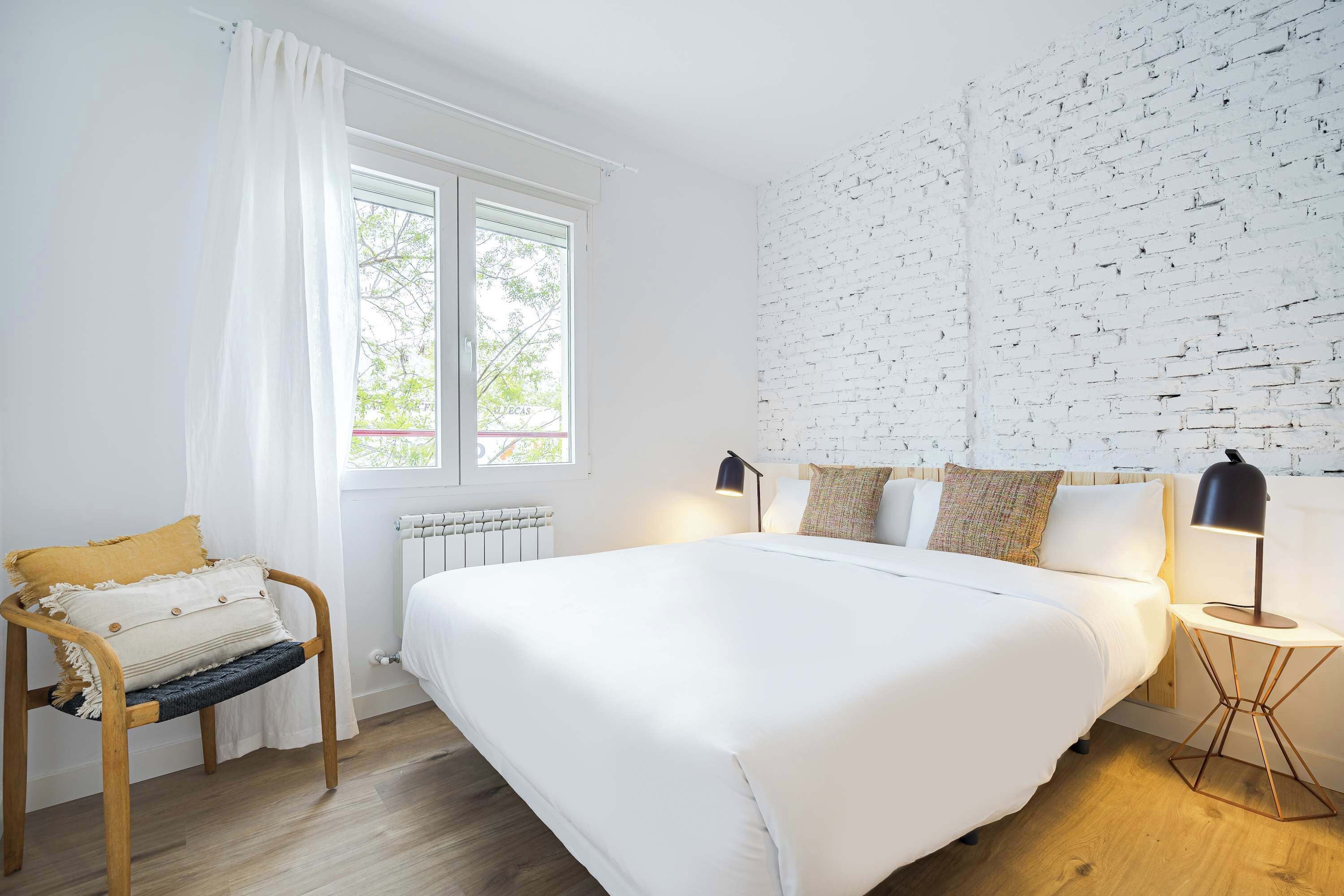 A bedroom with a large bed, a chair with cushions, a bedside table with a lamp, and a brick-patterned wall.