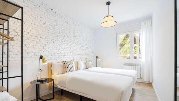 A modern bedroom with two beds, a white brick wall, a wooden floor, and a window with curtains. - Vallecano Apartments by Olala Homes (Madrid)