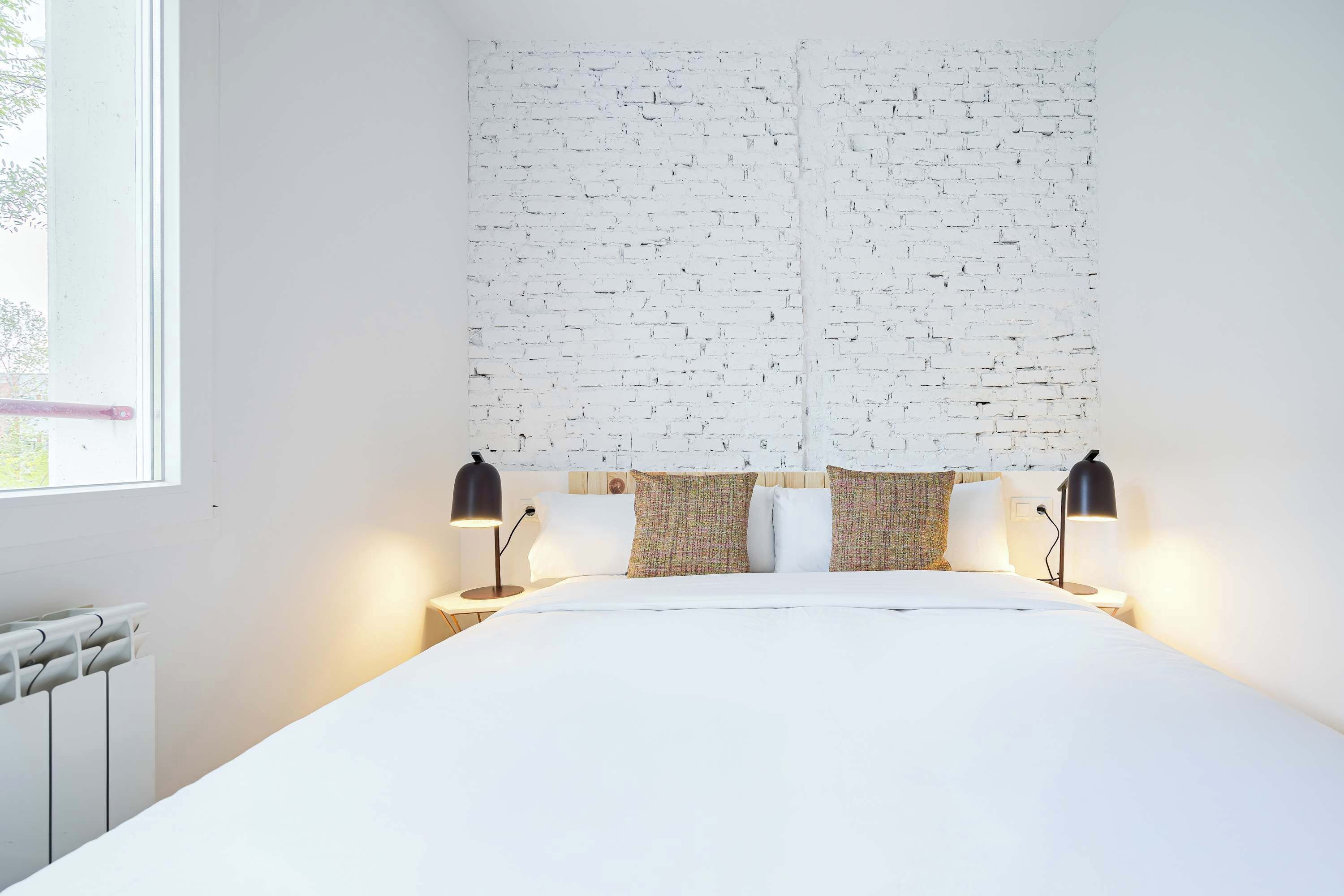 Ein modernes Hotelzimmer mit weißer Ziegelwand, einem großen Bett, zwei Nachttischlampen und einem Fenster mit Blick ins Grüne.