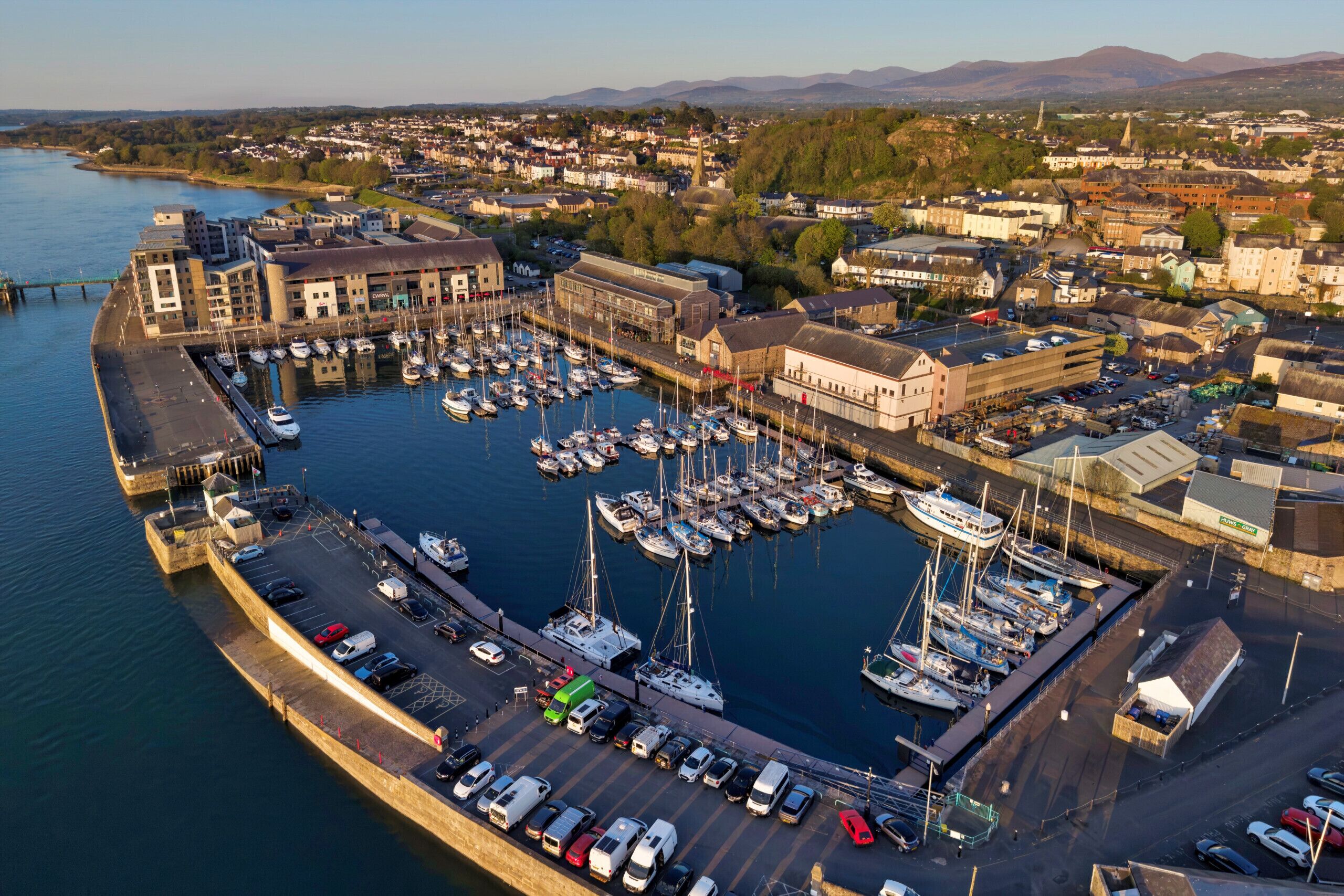 An aerial view of a marina with boats docked, a parking area, and surrounding buildings.