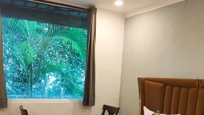 A bedroom with a bed, a window with a view of greenery, a ceiling fan, and a bedside table. - Haveli NH 8 (New Delhi)
