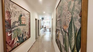 A hallway with two large floral paintings on the walls, a polished floor, and a view into a dining area with tables and chairs. - Haveli NH 8 (New Delhi)