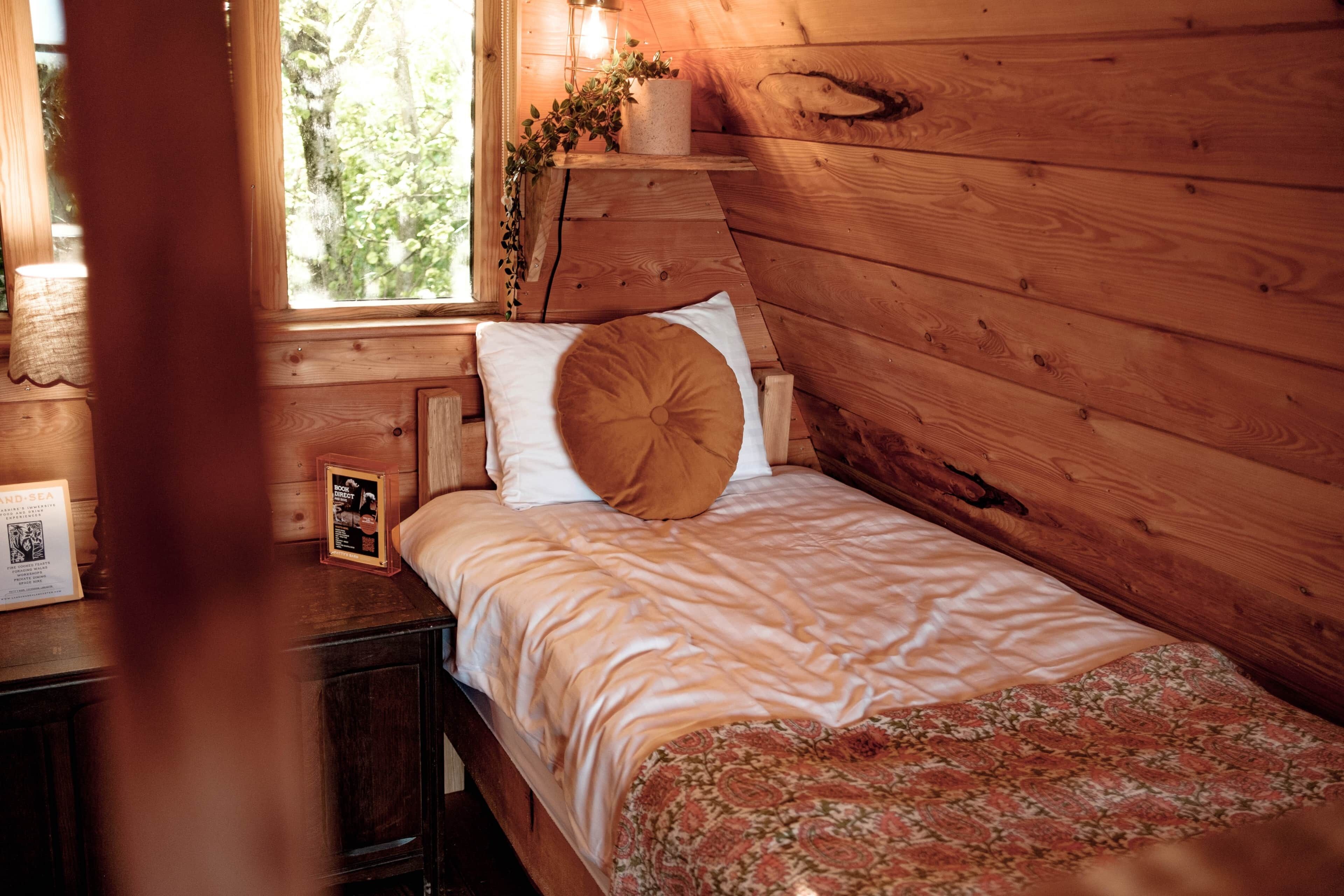 Ein Zimmer in einer Holzhütte mit einem Bett, einem Nachttisch, einem gerahmten Bild und einem Fenster mit Blick auf Grünflächen.