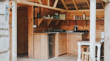 A wooden cabin with a kitchen area featuring a refrigerator, a coffee maker, and shelves with various items.
