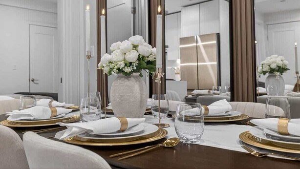 Una mesa de comedor puesta con servilletas blancas, cubiertos dorados y un florero con flores blancas.