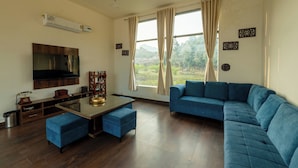 A living room with a blue sofa, a coffee table, a TV, and a view of greenery outside.