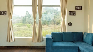 A room with a blue sofa, beige curtains, and a view of a green landscape through a large window.