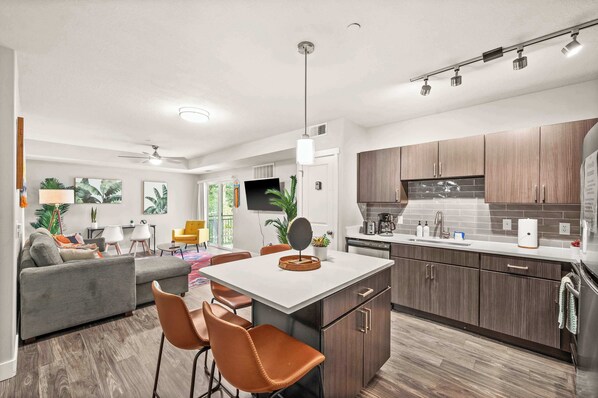 A modern kitchen with a central island, wooden cabinets, and a dining area with a grey sofa and orange chairs.
