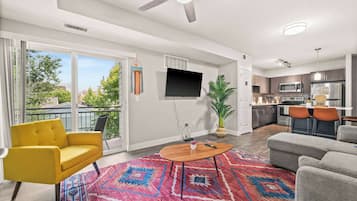 Un salon moderne avec un canapé jaune, un canapé gris, une table basse en bois et un tapis coloré. Un téléviseur à écran plat est fixé au mur, il y a un ventilateur de plafond et une grande fenêtre donnant sur des arbres et des immeubles.