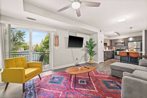 A modern living room with a yellow sofa, a gray sofa, a wooden coffee table, and a colorful rug. There is a flat-screen TV mounted on the wall, a ceiling fan, and a large window with a view of trees and buildings.