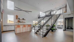 A spacious kitchen with a central island, a staircase with a metal railing, and a view of the city through large windows.