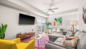 A modern living room with a grey sofa, yellow armchairs, a flat-screen TV, a ceiling fan, and a large window with a view of the outdoors.