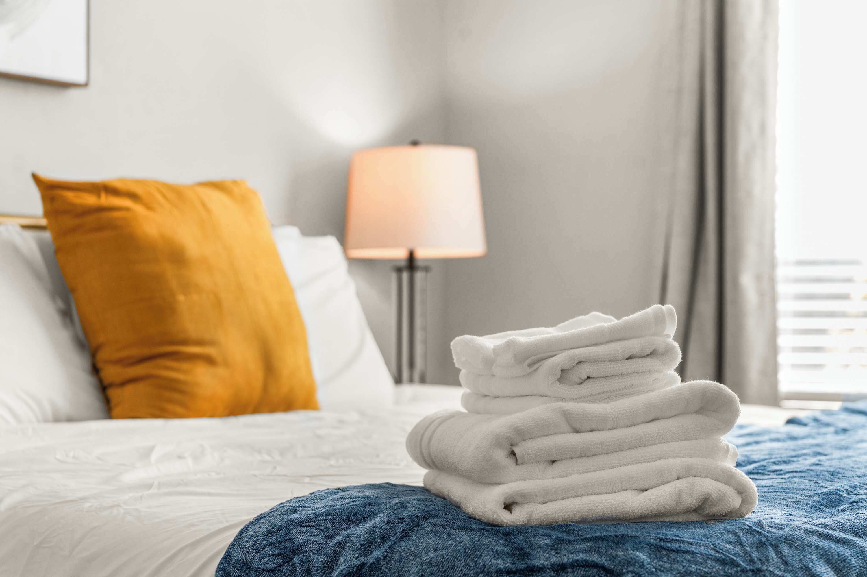 A neatly made bed with white and blue bedding, a stack of white towels, and a mustard-colored pillow.
