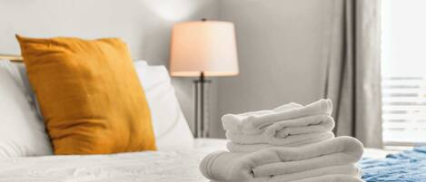 A neatly made bed with white and blue bedding, a stack of white towels, and a mustard-colored pillow.