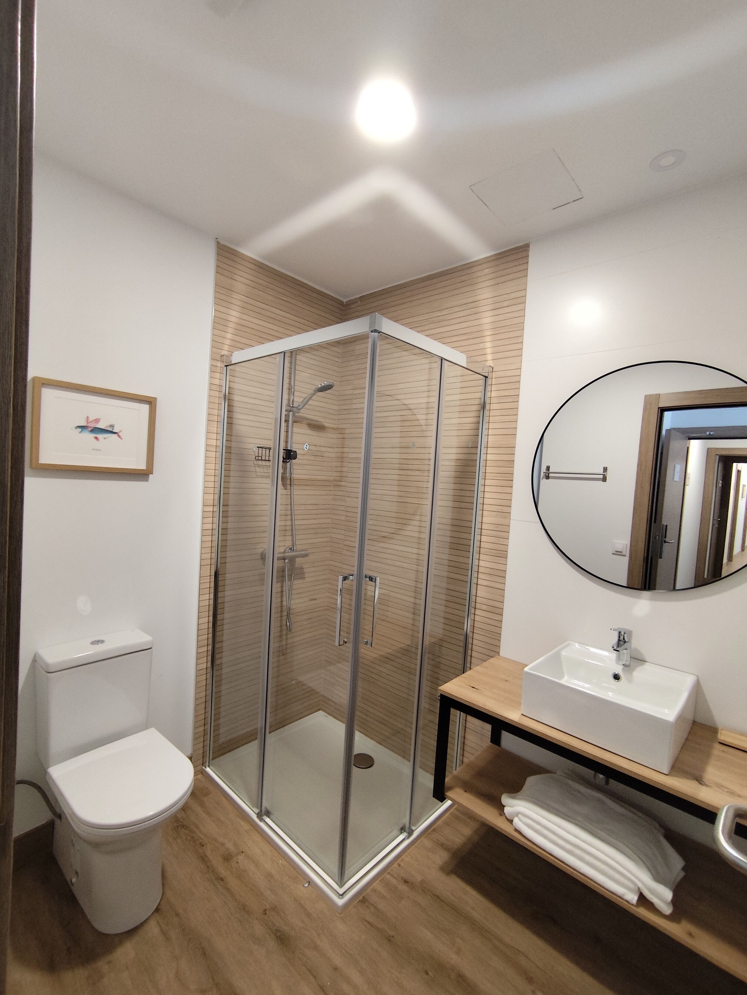 Egy modern fürőszoba, melyben üvegfalas zuhanyzó, WC, mosdókagyló és tükör található.