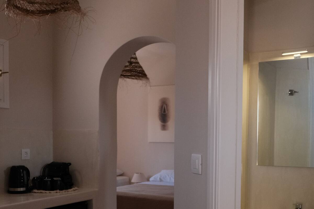 Habitación de hotel con kitchenette, baño con espejo y cama con cabecera blanca.
