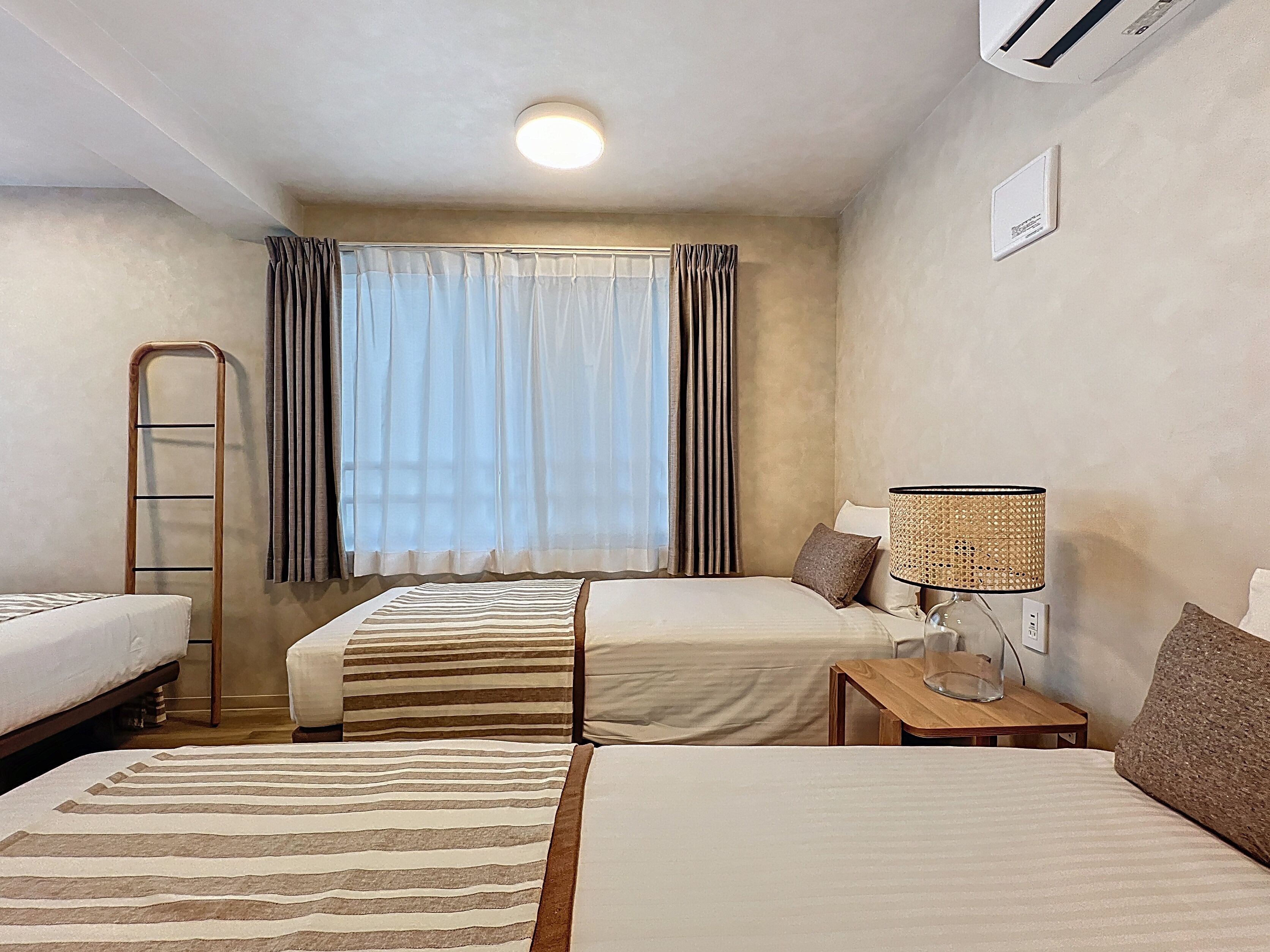 A hotel room with two beds, a window with curtains, a bedside table with a lamp, and a wall-mounted air conditioner.