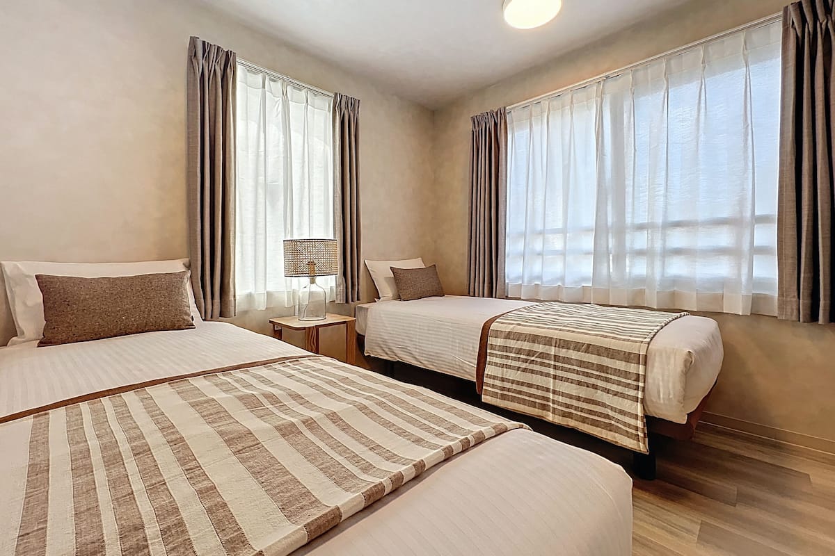 A hotel room with two beds, a bedside table with a lamp, and large windows with curtains.