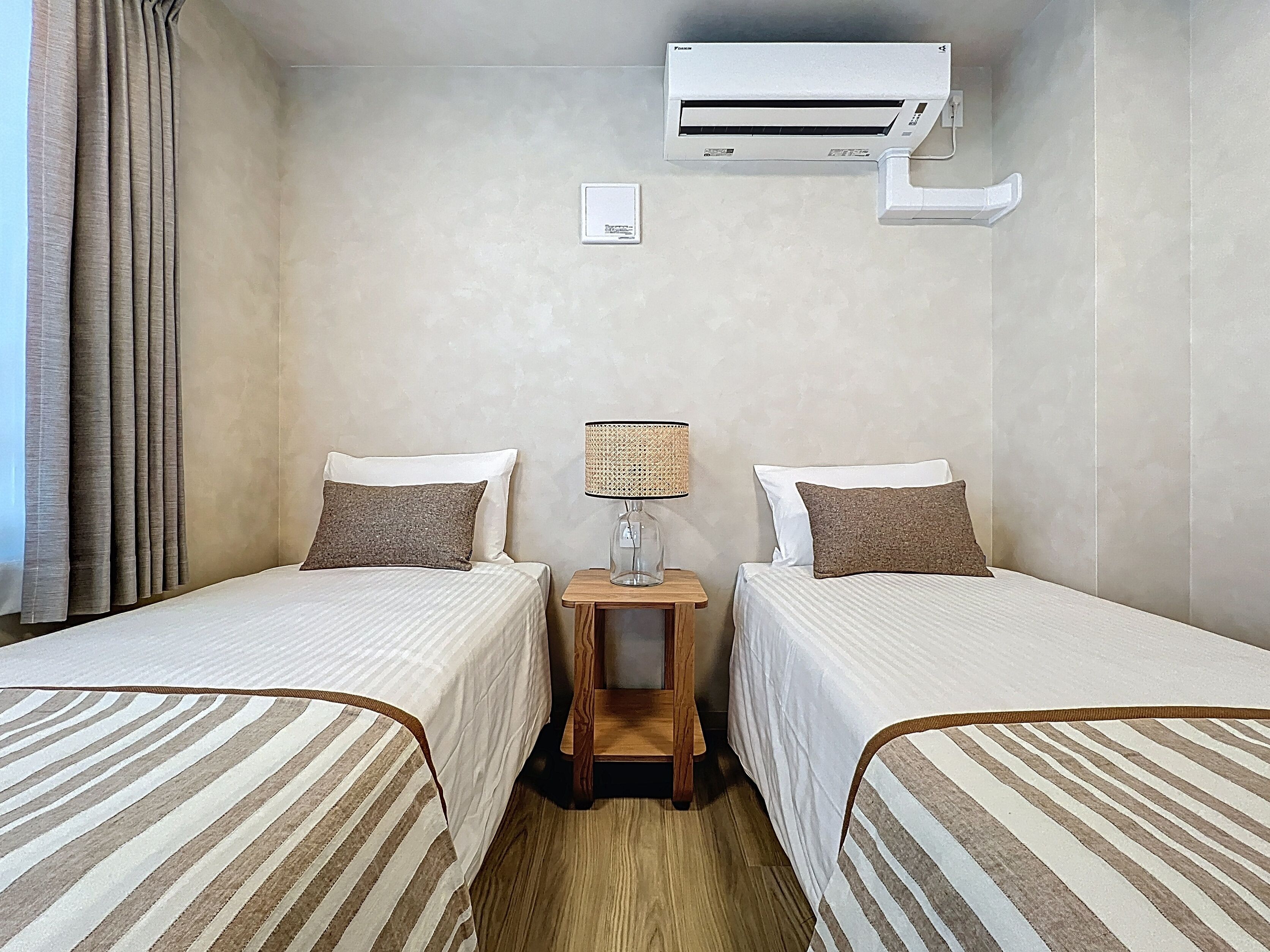 A hotel room with two beds, a nightstand with a lamp, an air conditioning unit, and a window with curtains.