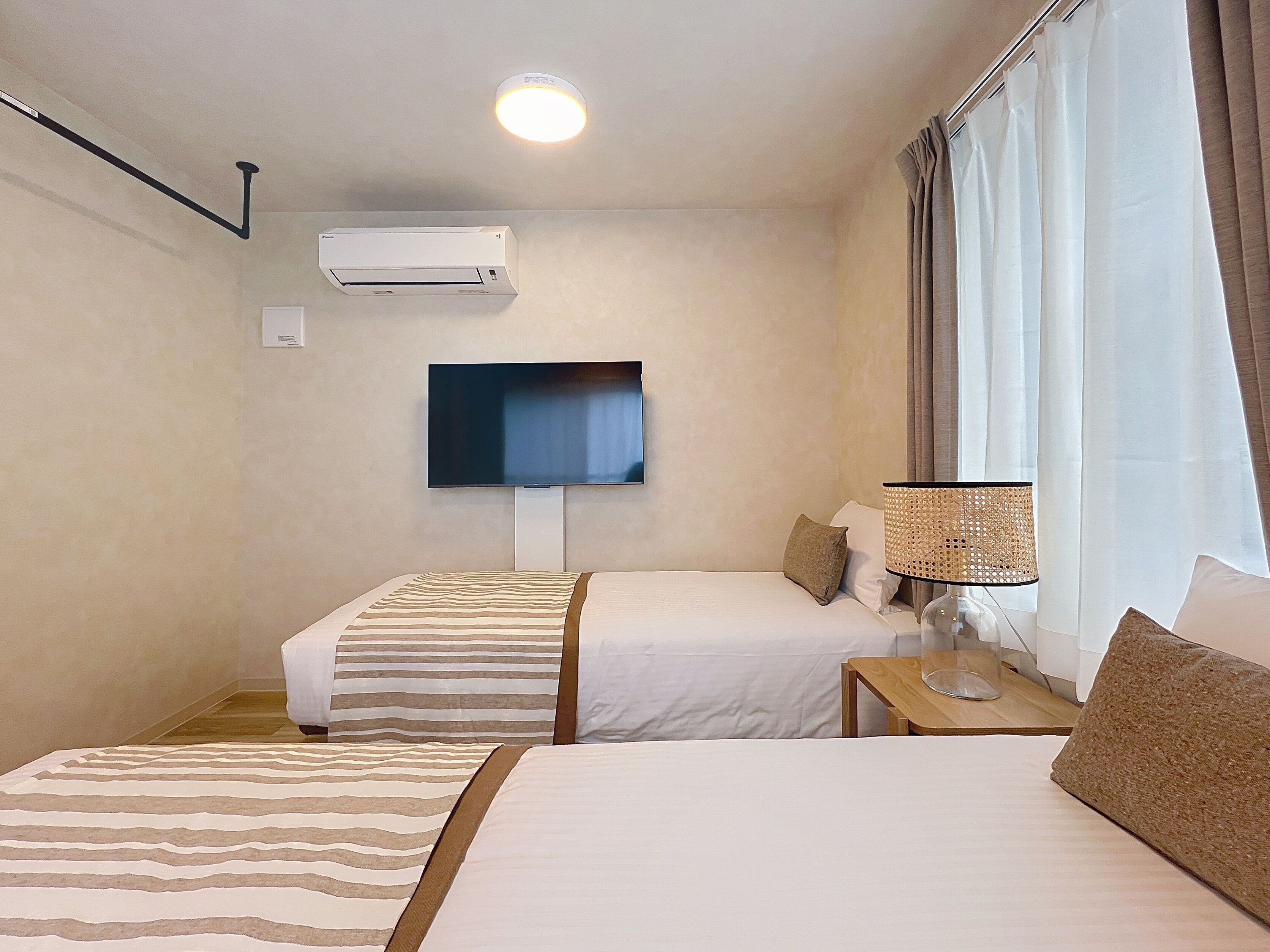 Habitación de hotel con dos camas, un televisor de pantalla plana montado en la pared, un aire acondicionado, una mesita de noche con lámpara y una ventana con cortinas.