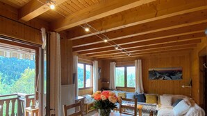 A wooden cabin interior with a dining table, chairs, a sofa, and a view of the forest through large windows. - Chalet on the edge of town, close to the Balme cable car (La Clusaz)