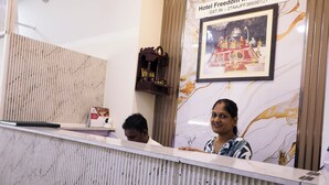 Bancada de recepção de hotel com uma moldura com imagem de um templo, uma prateleira de madeira com objetos, e uma placa com o nome do hotel e número GST.